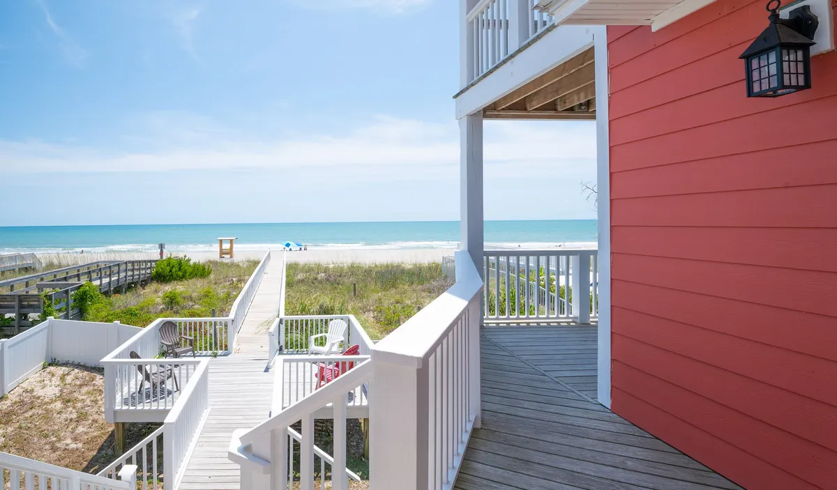 The image showcases a classic red oceanfront house with expansive wraparound decks, a private boardwalk leading directly onto the soft sands of Kure Beach, and the sparkling Atlantic stretching out beyond. The scene captures the relaxed ambiance, with sunlight bathing the porch and beach, inviting guests to unwind, gather with family and friends, or step onto the sand for early morning yoga or evening strolls.