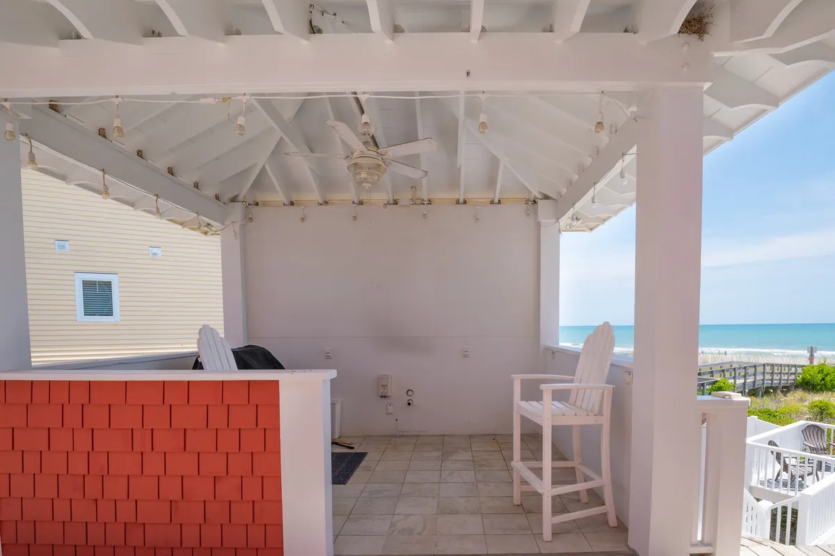 An open patio area featuring white wooden chairs, a ceiling fan, and a stunning view of the ocean. The inviting setup suggests a tranquil spot for relaxing, reading, or spending time with friends and pets while enjoying the seaside breeze.
