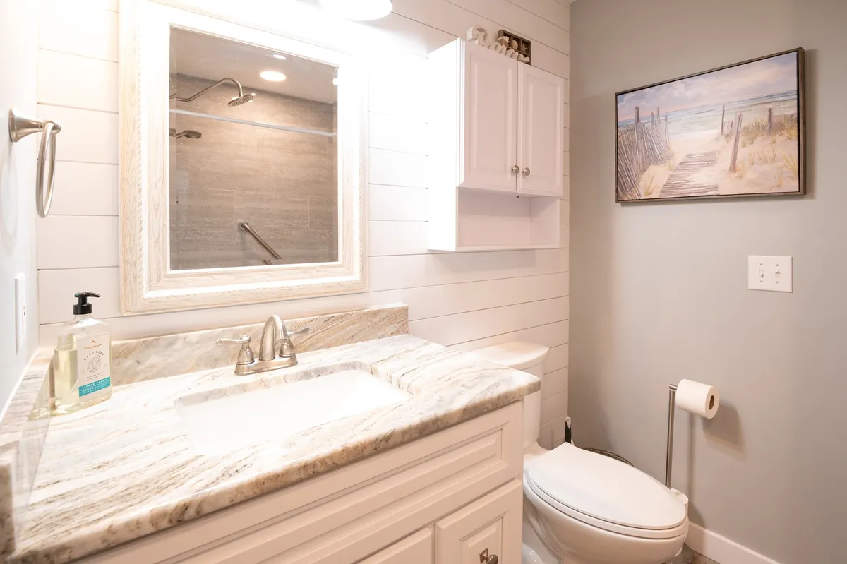 A remodeled bathroom featuring a sleek granite countertop, a spacious mirror, shower area, and tasteful wall art, all bathed in refreshing natural light. The space looks recently updated and inviting for morning or evening routines.