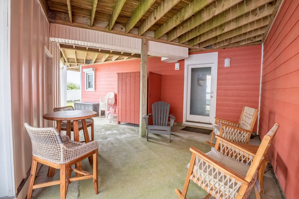 A cozy outdoor space features a wooden table and chairs, colorful walls, a red storage shed, and classic rocking chairs, set up for relaxation and socializing. The open, casual environment evokes the idea of casual drinks and conversations in a setting that transitions easily between indoor and outdoor living.