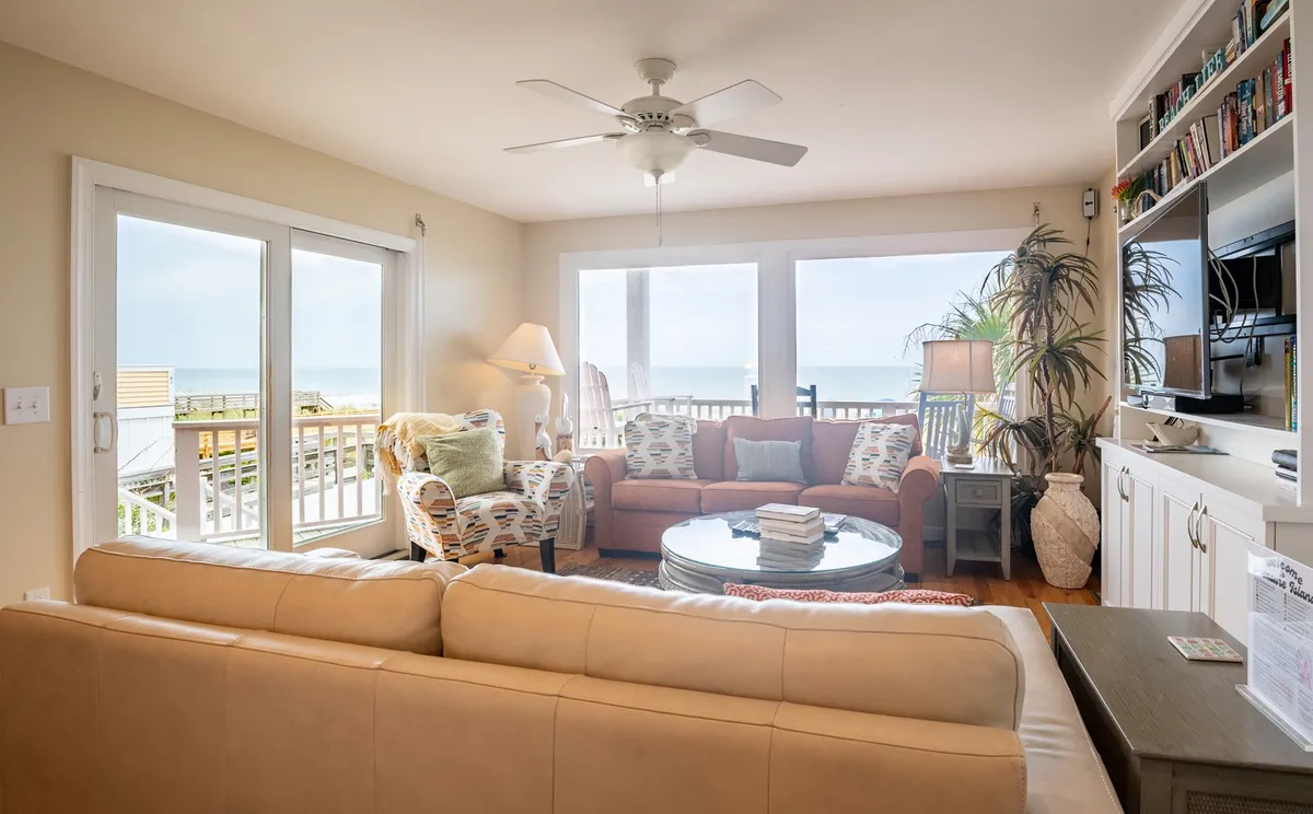 A comfortable seating area inside a beach house, with large windows offering an ocean view. Plush seating is arranged for casual conversation or relaxation, and a coffee table with books invites guests to unwind while enjoying the scenery. The overall ambiance is cozy and welcoming, ideal for regrouping after a day of exploring.