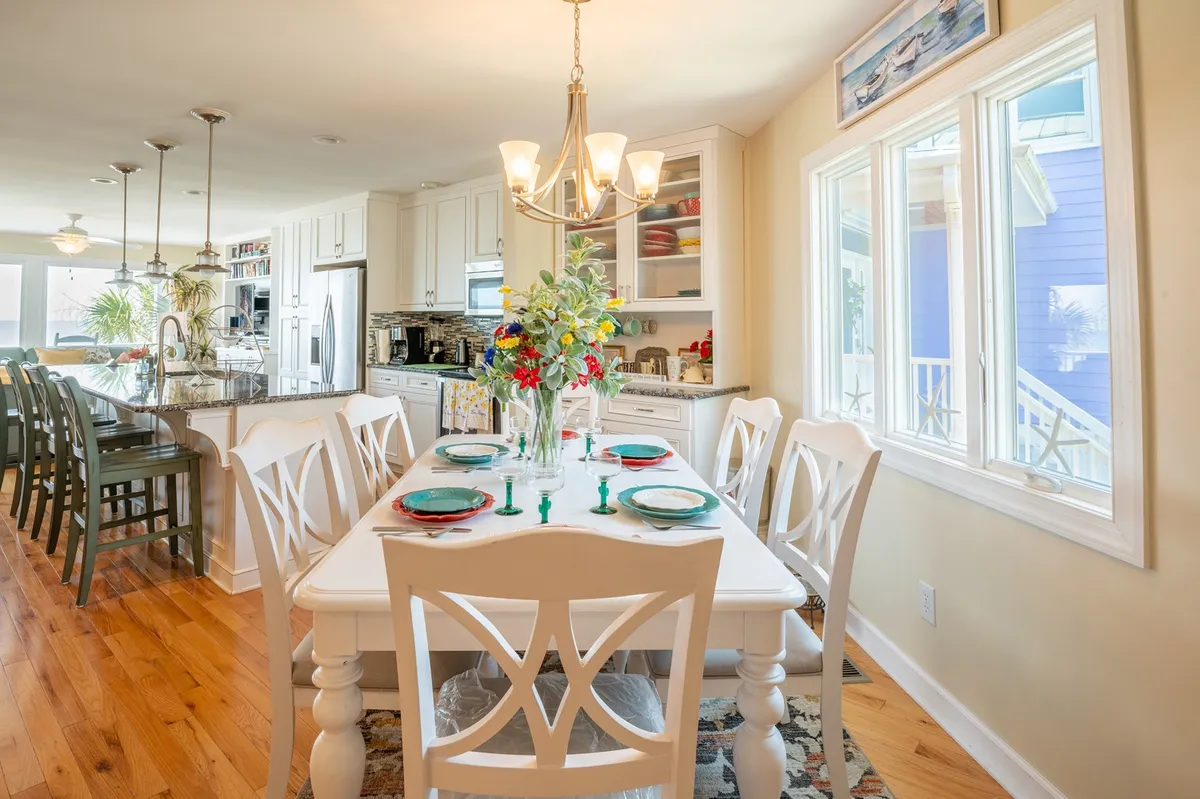 A bright, airy dining table with colorful place settings and a floral centerpiece sits in front of large windows that flood the space with natural light. The atmosphere is vibrant, warm, and inviting, designed for memorable meals and celebrations.