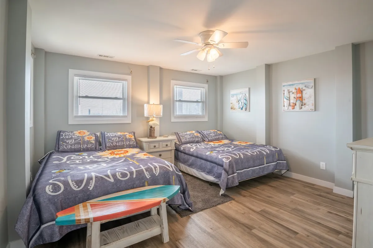 A playful yet comfortable bedroom with two double beds adorned in sunny-themed bedding, surfboard decor on the wall, and a nightstand with a lamp. The arrangement and decor suggest a family- or kid-friendly space with a relaxed, coastal vibe.