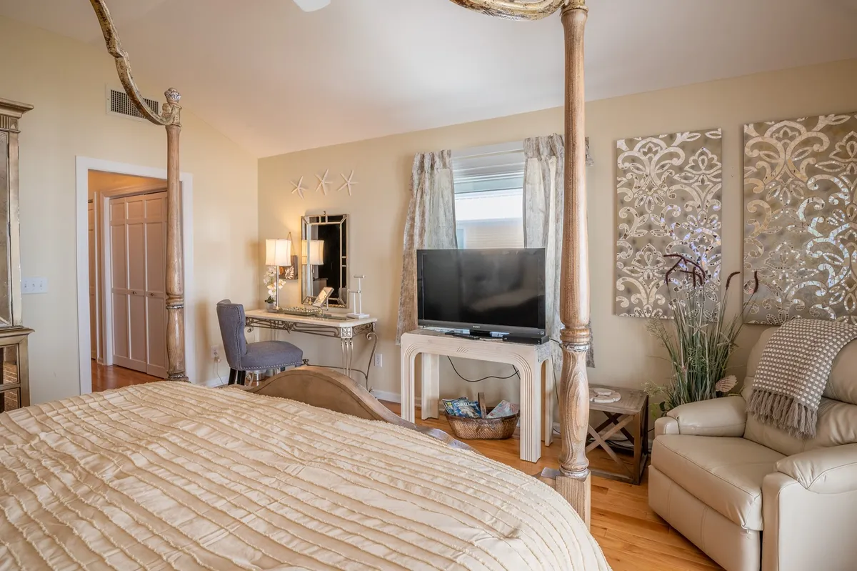 A spacious and comfortable bedroom with a canopy bed, decorative wall panels, a television, an armchair, and a dressing table. Soft natural lighting enhances the serene mood, making it ideal for both relaxation and quiet entertainment.
