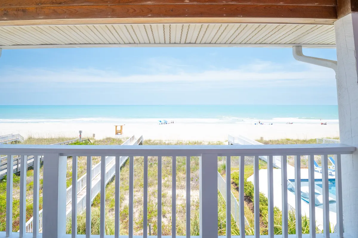 A wide, open view of the beach and ocean, seen from the end of a private boardwalk. Pristine white sand, gentle waves, and a clear blue sky invite exploration, while the wooden walkway draws the eye straight to the water's edge.