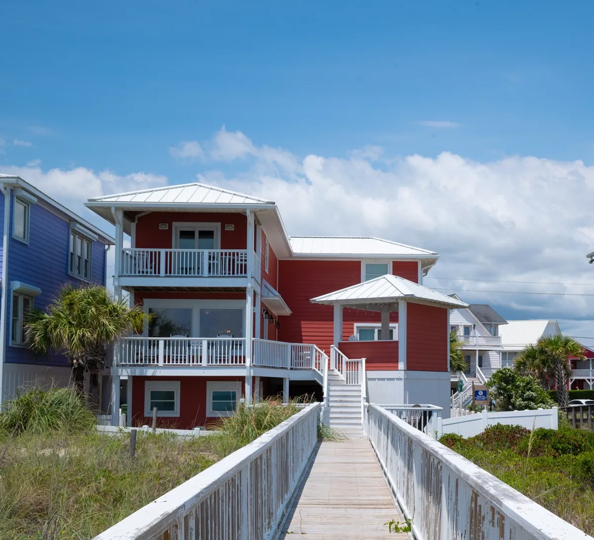This exterior image captures a cheerful red beach house surrounded by lush greenery. A private wooden walkway leads from the home toward the beach, evoking a strong sense of direct beach access and outdoor living. The home’s multiple decks and wraparound porch are visible, with seating arranged for enjoying the outdoors, sunsets, or meals together.