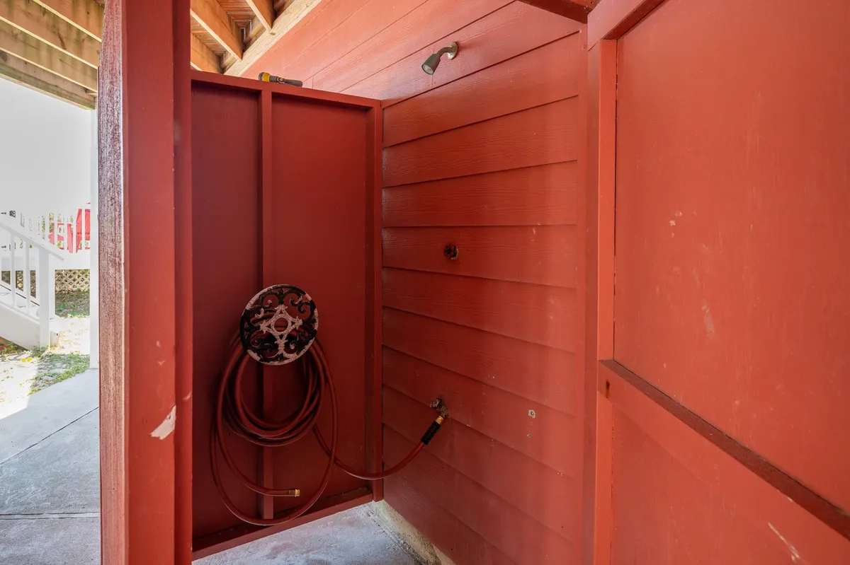 A practical and stylish red outdoor shower enclosure with a hose reel, set against wooden paneling. The space is designed for rinsing off after beach outings, capturing the transition from sandy adventures to relaxed comfort.