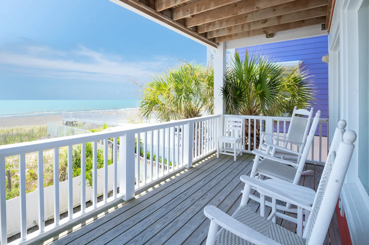 A tranquil exterior scene featuring classic rocking chairs on a sun deck, framed by palm trees and offering a panoramic ocean view. Early morning sunlight reflects off the water, creating an inviting atmosphere for enjoying ocean breezes and peaceful moments.
