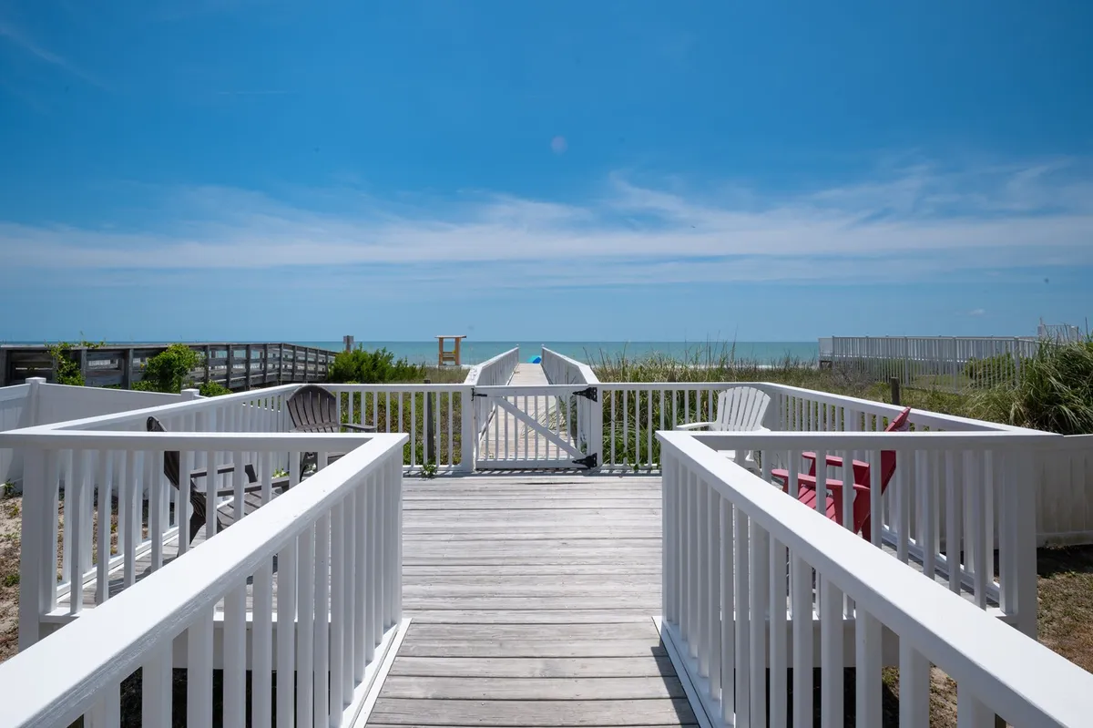 A tranquil scene featuring a wooden boardwalk stretching towards the sandy beach, with beach chairs set up to enjoy the vast ocean views. The open sky and gentle waves suggest endless opportunities for sunbathing, swimming, or strolls along the shore.