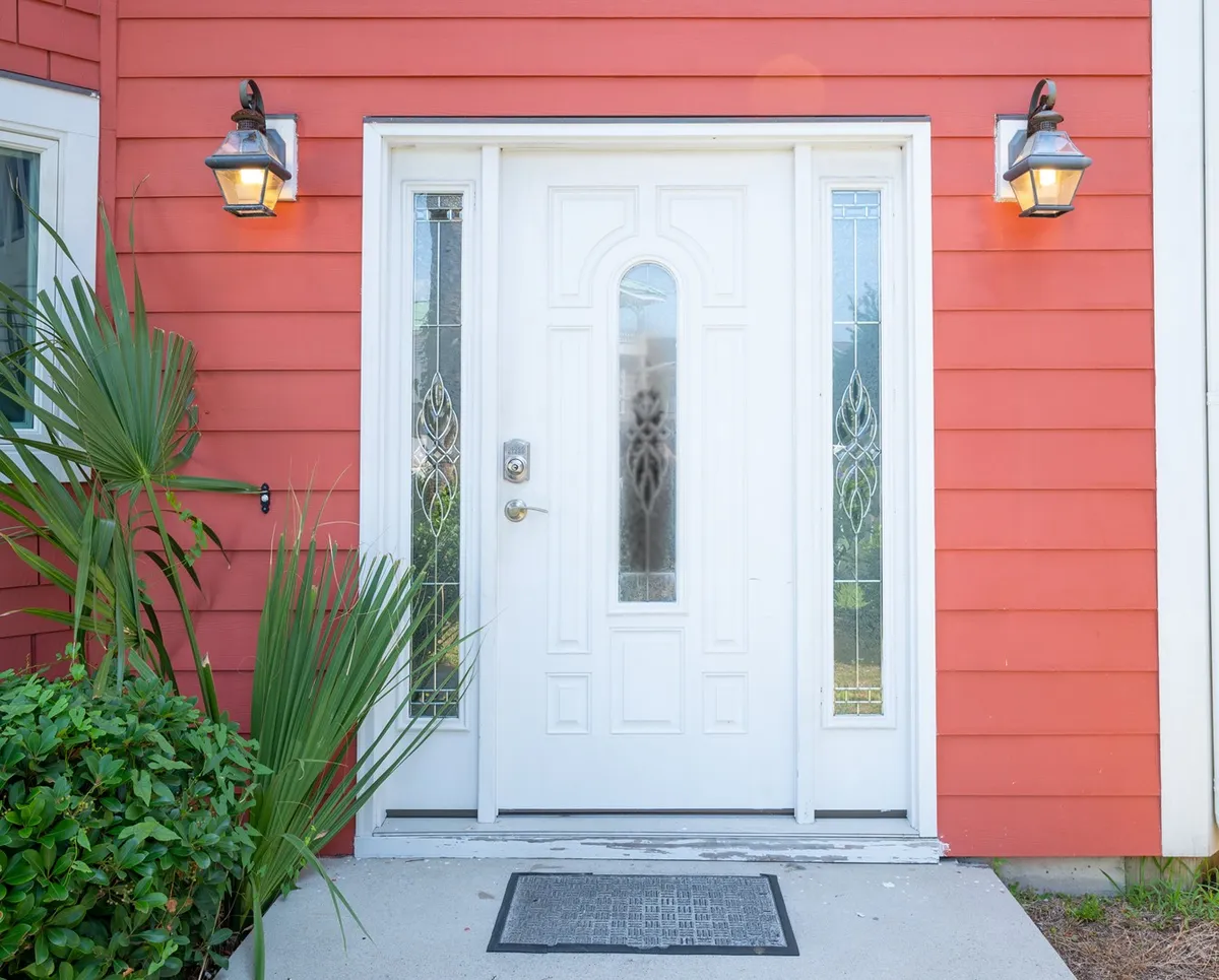 An inviting entryway with a white door featuring decorative glass panels, set in red siding and surrounded by green plants. The porch area hints at a warm welcome and community feel.