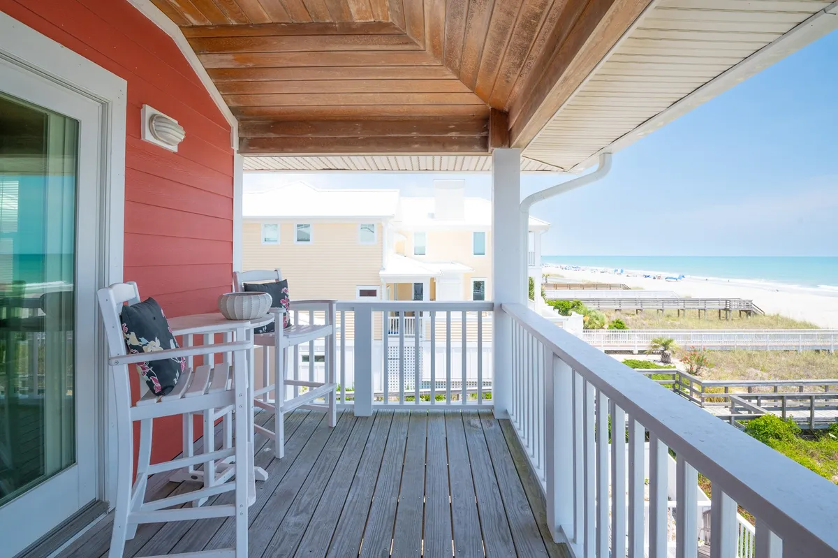 An exterior view showcasing a charming red wall with white railing, stylish outdoor seating, and a sweeping beach view beyond. The setting evokes a seamless connection to the ocean and provides a welcoming outdoor environment.